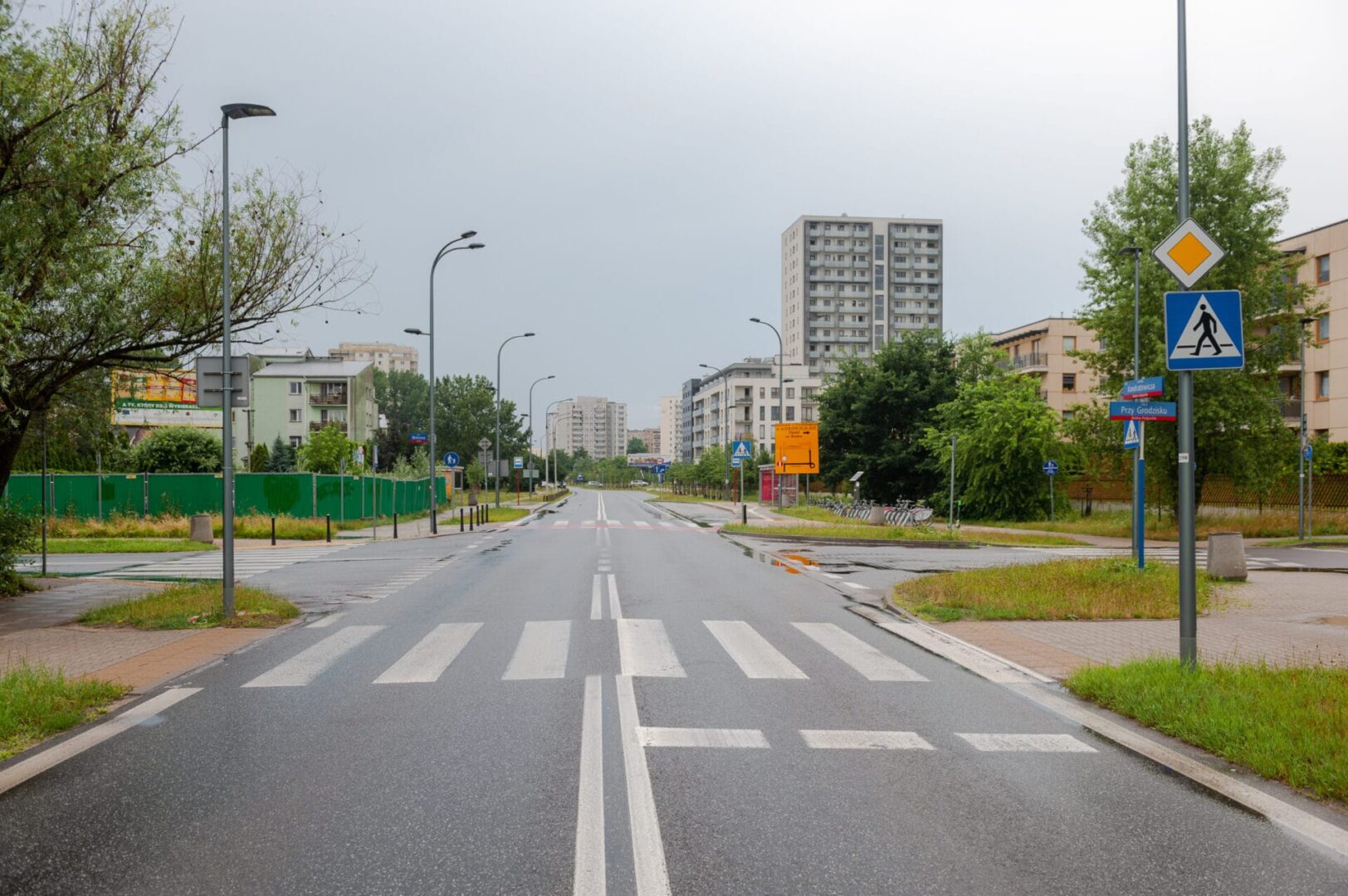Pusta ulica miejska z przejściem dla pieszych, budynkami mieszkalnymi i latarniami ulicznymi.