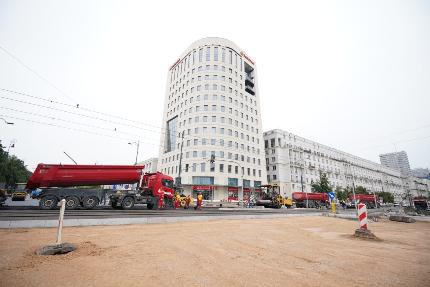 Budowa drogi w pobliżu budynku Santander Bank. Widoczne wywrotki i sprzęt do układania nawierzchni.