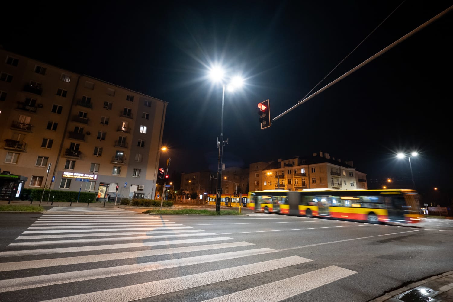 Nocna scena uliczna z przejściem dla pieszych, autobusami i sygnalizacją świetlną.