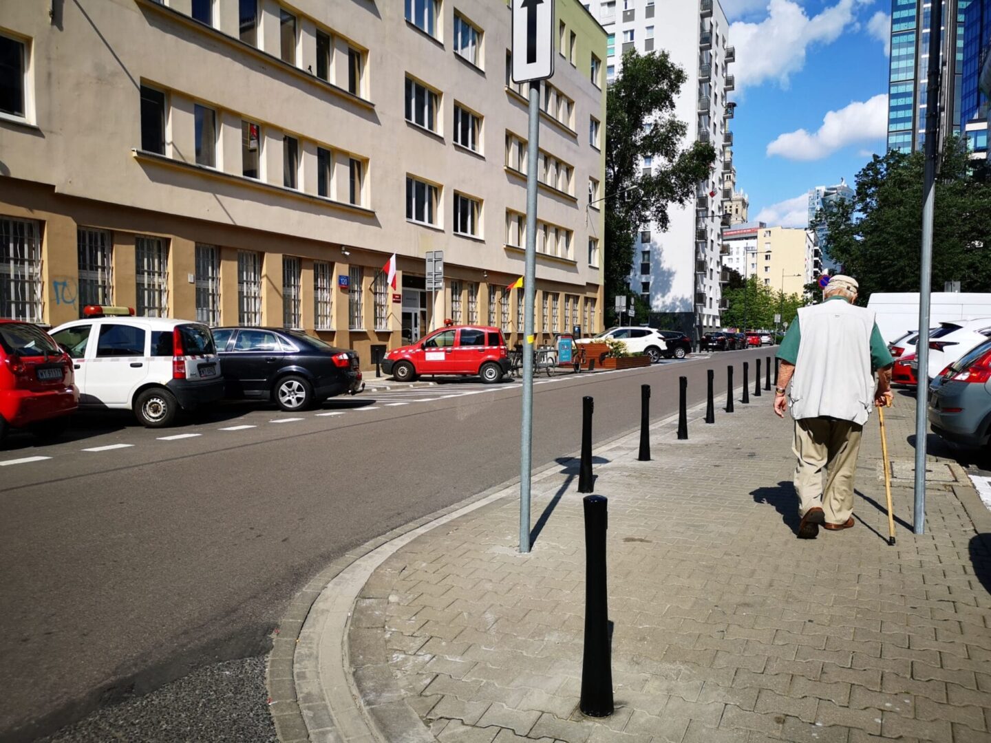 Starszy mężczyzna idzie o lasce obok zaparkowanych samochodów na ulicy miejskiej.