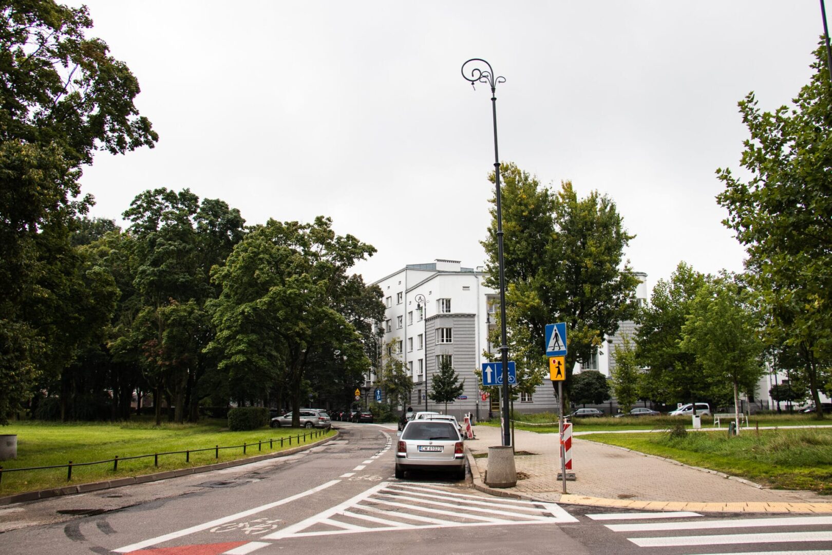 Scena uliczna z zaparkowanymi samochodami, drzewami i nowoczesnym budynkiem mieszkalnym.