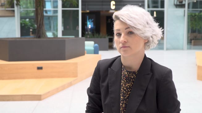 Młoda kobieta z krótkimi, białymi włosami, ubrana w czarny żakiet, siada do rozmowy kwalifikacyjnej.