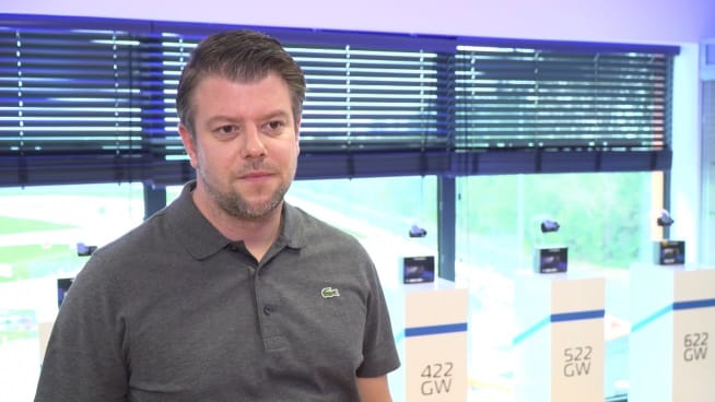 Mężczyzna w szarej koszulce polo mówi, za nim eksponowane są produkty