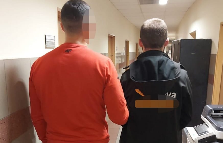 Dwóch mężczyzn idących korytarzem, jeden w pomarańczowej koszuli, drugi w czarnej kurtce.