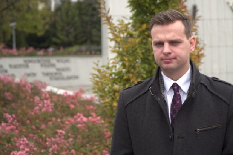 Mężczyzna w ciemnym płaszczu i krawacie, stojący na zewnątrz.