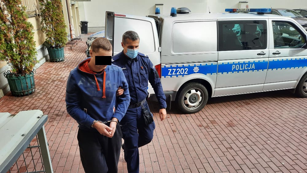 Policjant eskortujący podejrzanego w kajdankach w pobliżu radiowozu ZZ202