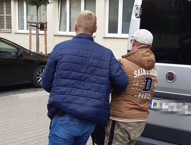 Dwóch mężczyzn w kajdankach eskortowanych przez policję do furgonetki.