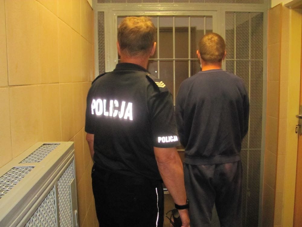 Policjant eskortujący więźnia na korytarzu więzienia. Na mundurze policjanta widoczny jest napis POLICJA.