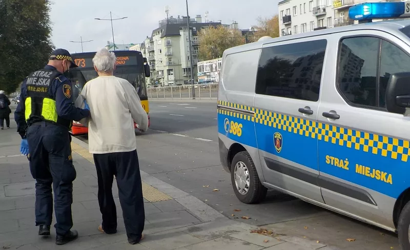 Policjant pomagający starszemu mężczyźnie w pobliżu autobusu miejskiego i furgonetki patrolowej. Straż Miejska.