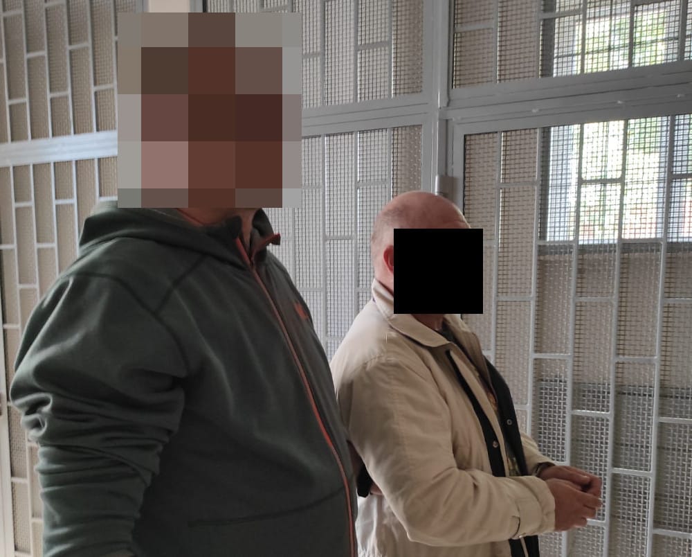 Dwóch mężczyzn w kajdankach stoi przed metalową siatką na ścianie.