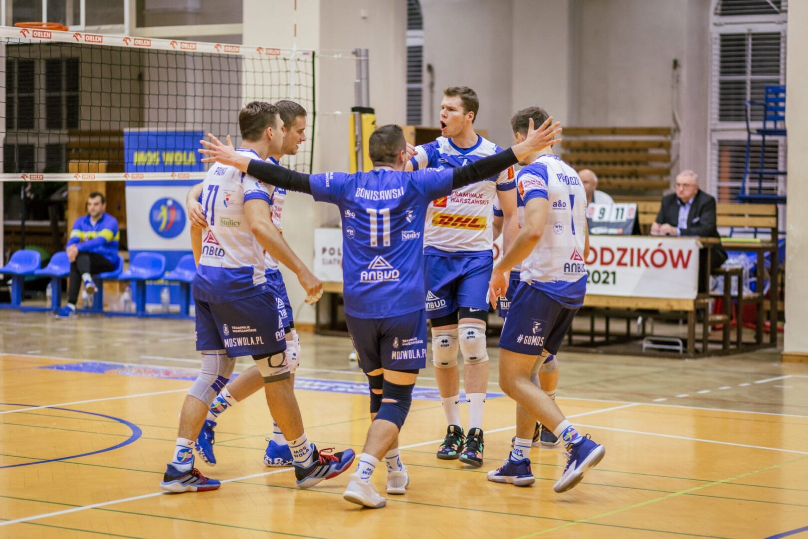 Siatkarze świętujący punkt, ubrani w niebiesko-białe stroje. Drużyna nazywa się MOS WOLA.