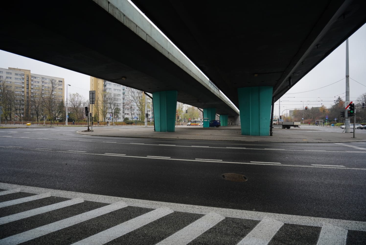 Pusta ulica miejska pod wiaduktem autostradowym, z turkusowymi filarami podtrzymującymi.
