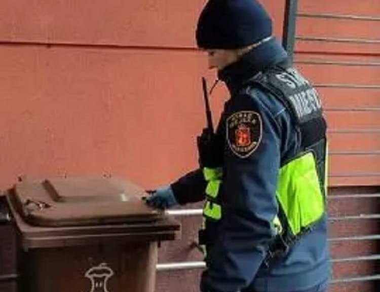 Policjant sprawdza brązowy, publiczny pojemnik na śmieci.