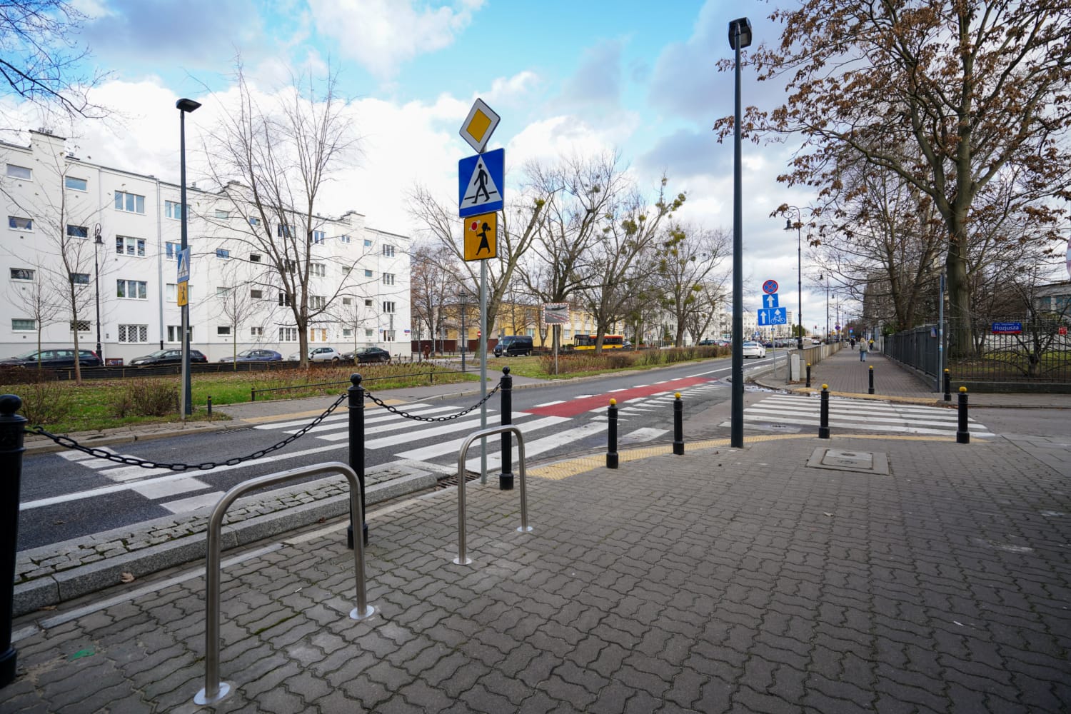 Przejście dla pieszych ze stojakami na rowery i znakami drogowymi na terenie osiedla mieszkaniowego.