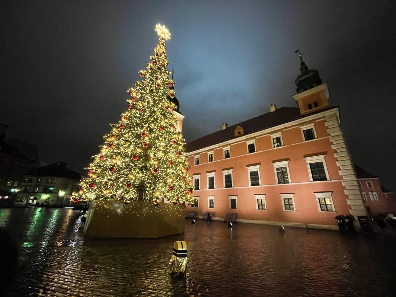 Duża choinka rozświetlona nocą na warszawskim Starym Mieście, obok zabytkowego budynku.