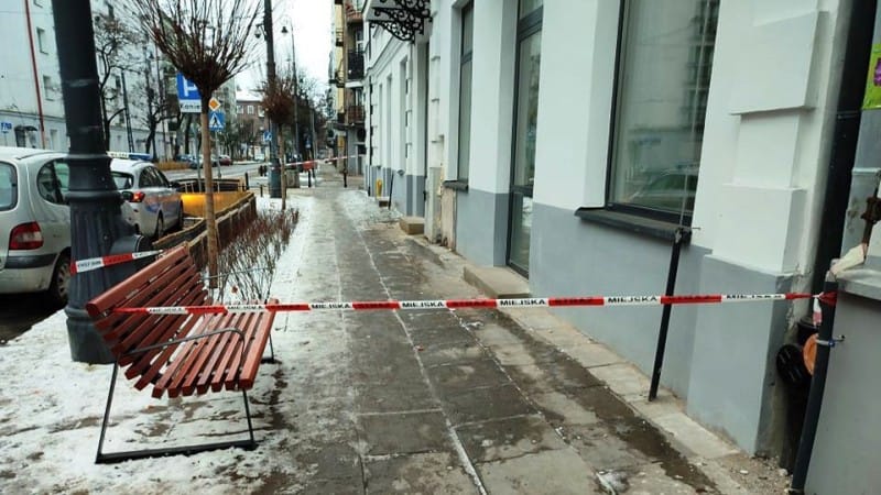 Taśma policyjna rozciągnięta na zaśnieżonym chodniku w pobliżu zaparkowanego samochodu i ławki.