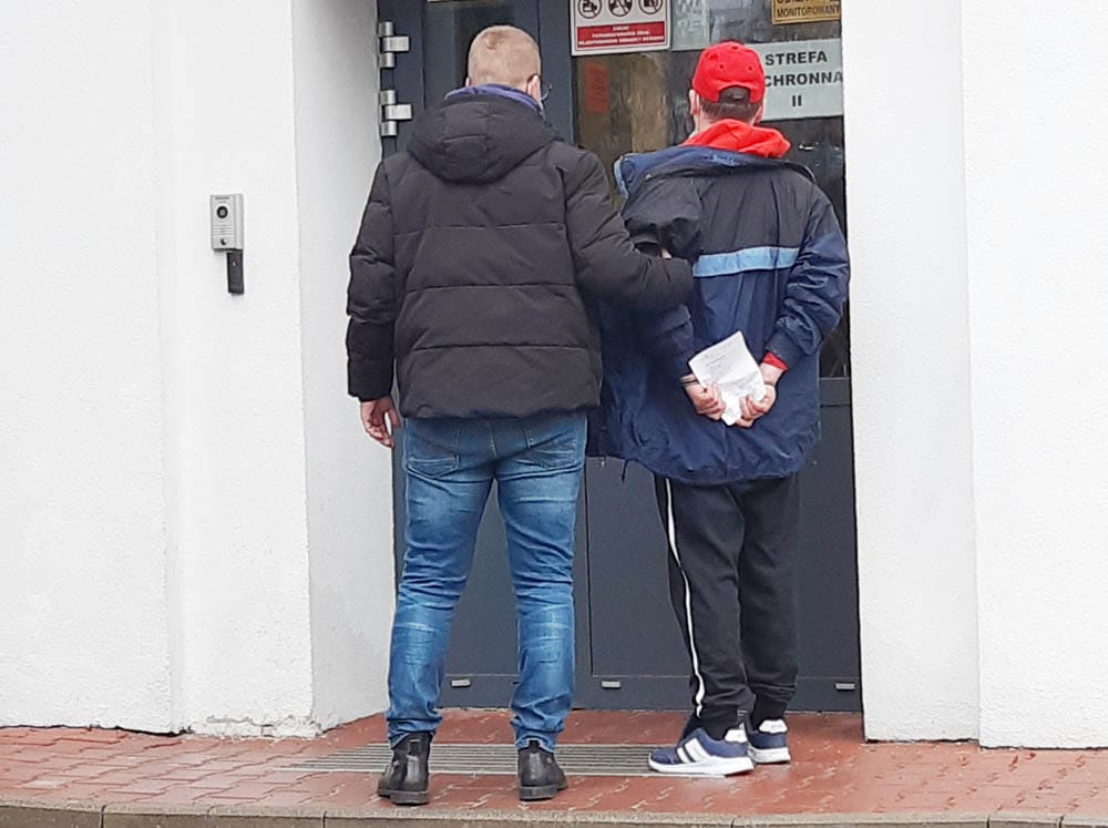 Dwóch mężczyzn stoi na zewnątrz budynku, jeden trzyma drugiego za ramię. Widoczna jest czerwona czapka.