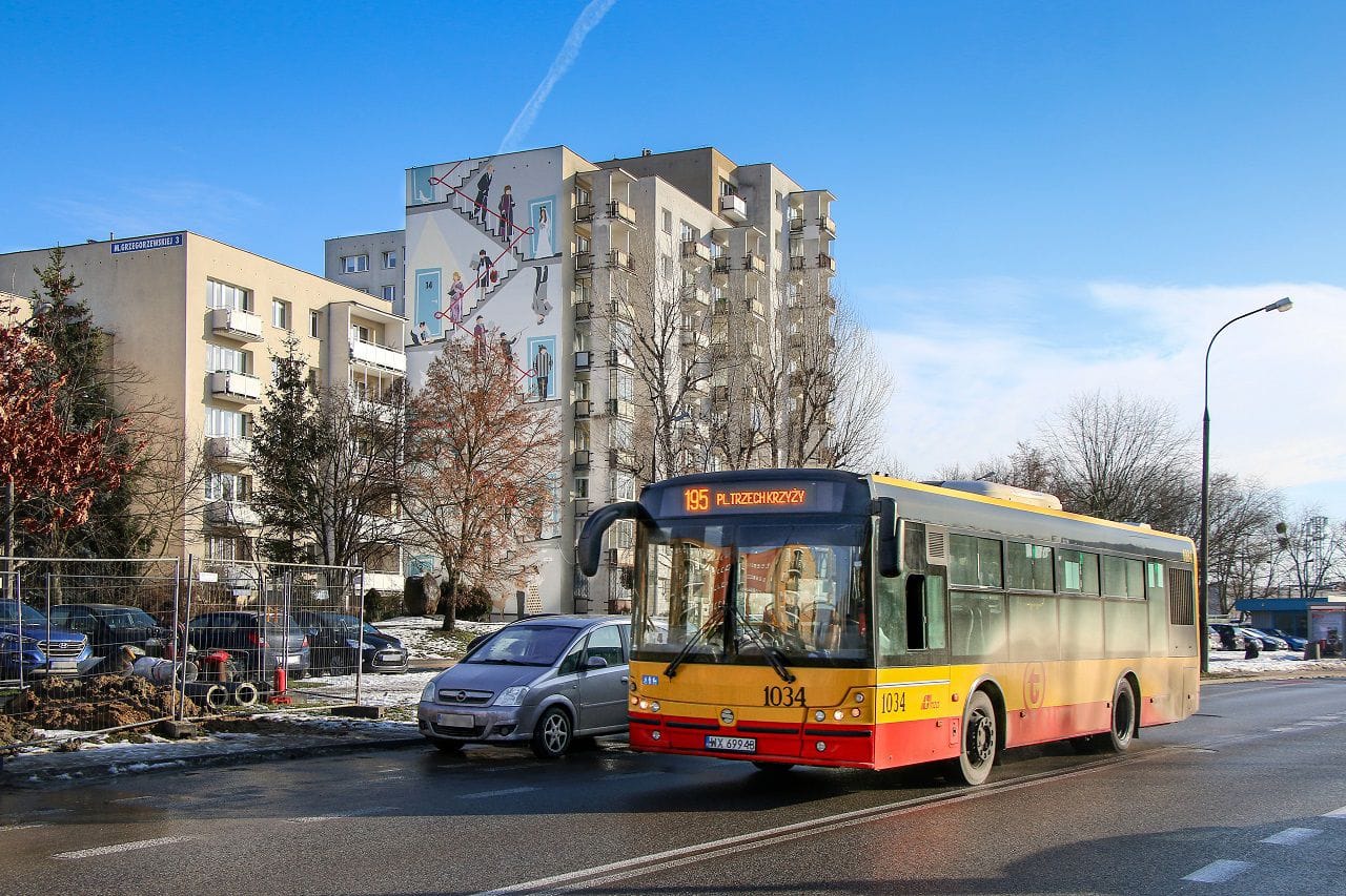 Autobus miejski 1034 na linii 195 do placu Trzech Krzyży, zaparkowany w pobliżu budynku z unikalnym muralem.