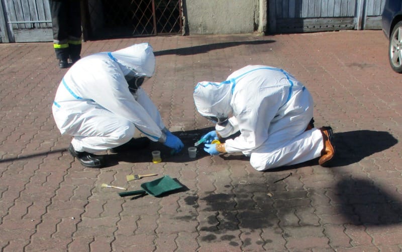 Dwóch śledczych ubranych w kombinezony ochronne zbiera dowody na miejscu zbrodni.