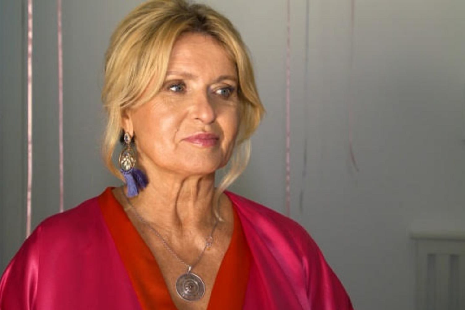 Dojrzała kobieta o blond włosach, ubrana w czerwony jedwabny szlafrok i z wyrazistą biżuterią.
