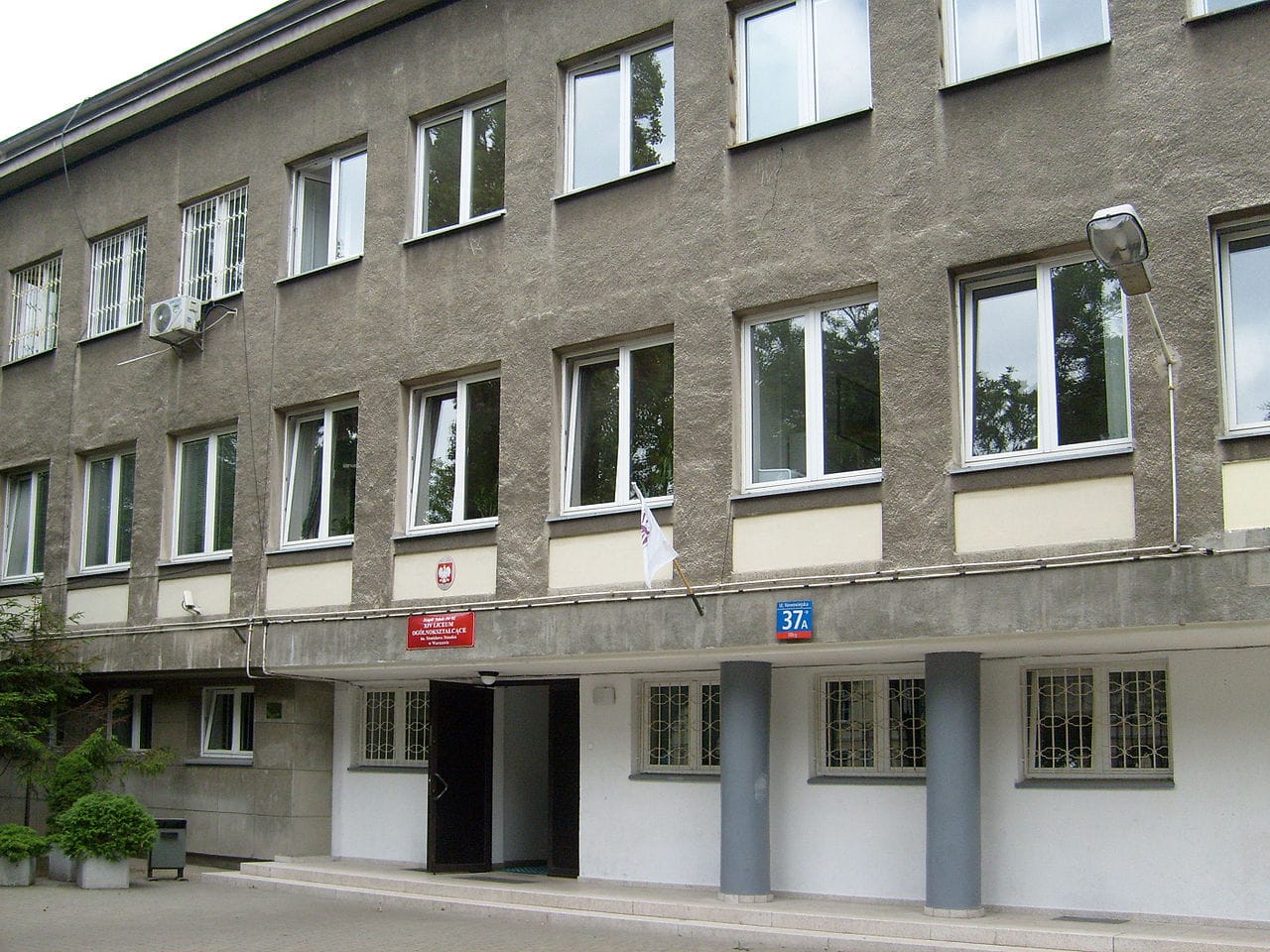 Zewnętrzna część szarego budynku z wieloma oknami, polską flagą i numerem 37A.