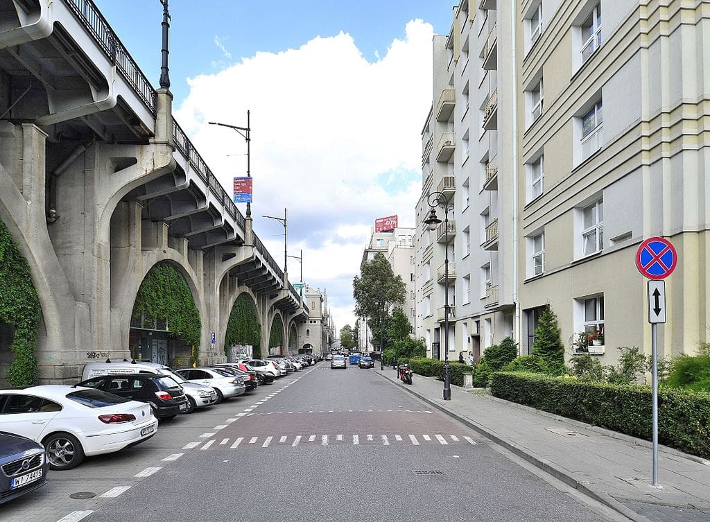 Scena uliczna w Warszawie. Samochody zaparkowane pod mostem, na tle nowoczesnych budynków mieszkalnych.