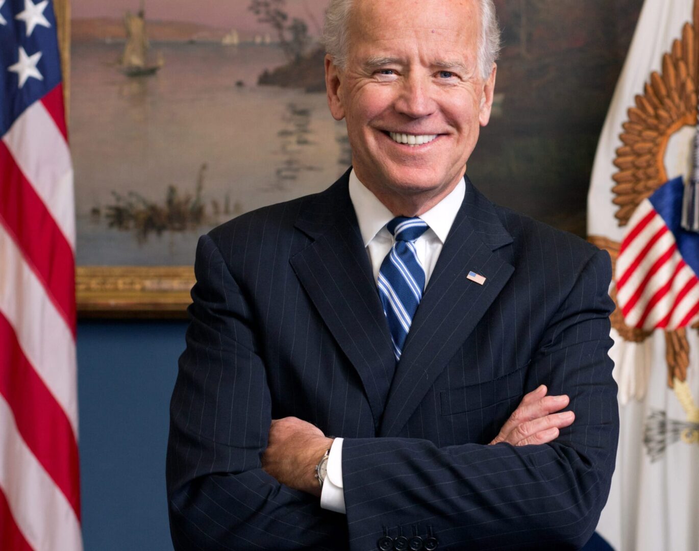 Prezydent Biden uśmiecha się i stoi ze skrzyżowanymi ramionami przed amerykańskimi flagami.