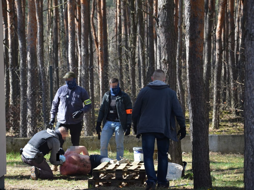 Policjanci badają miejsce zbrodni w zalesionym terenie. Ciało leży na ziemi.