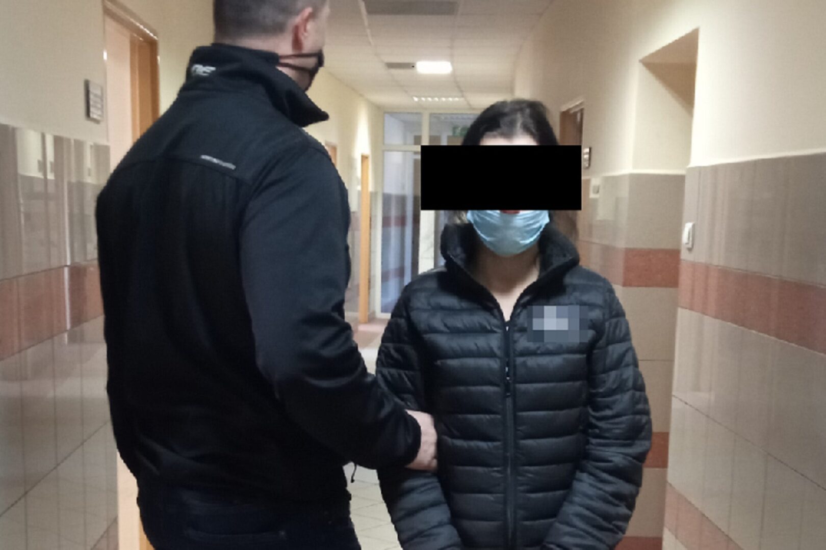 Policjant eskortujący zamaskowaną kobietę korytarzem.