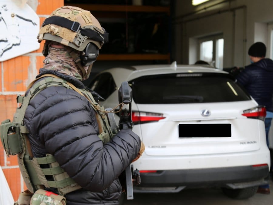 Funkcjonariusz SWAT z karabinem, w tle biały SUV Lexus
