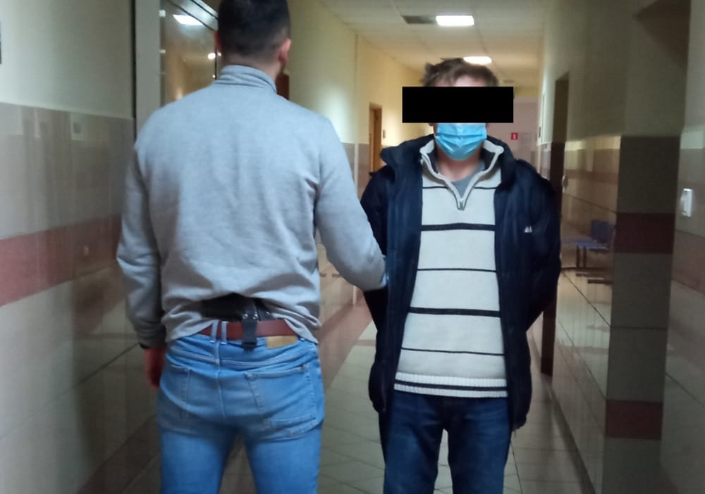 Policjant eskortujący zamaskowanego podejrzanego korytarzem szpitalnym.