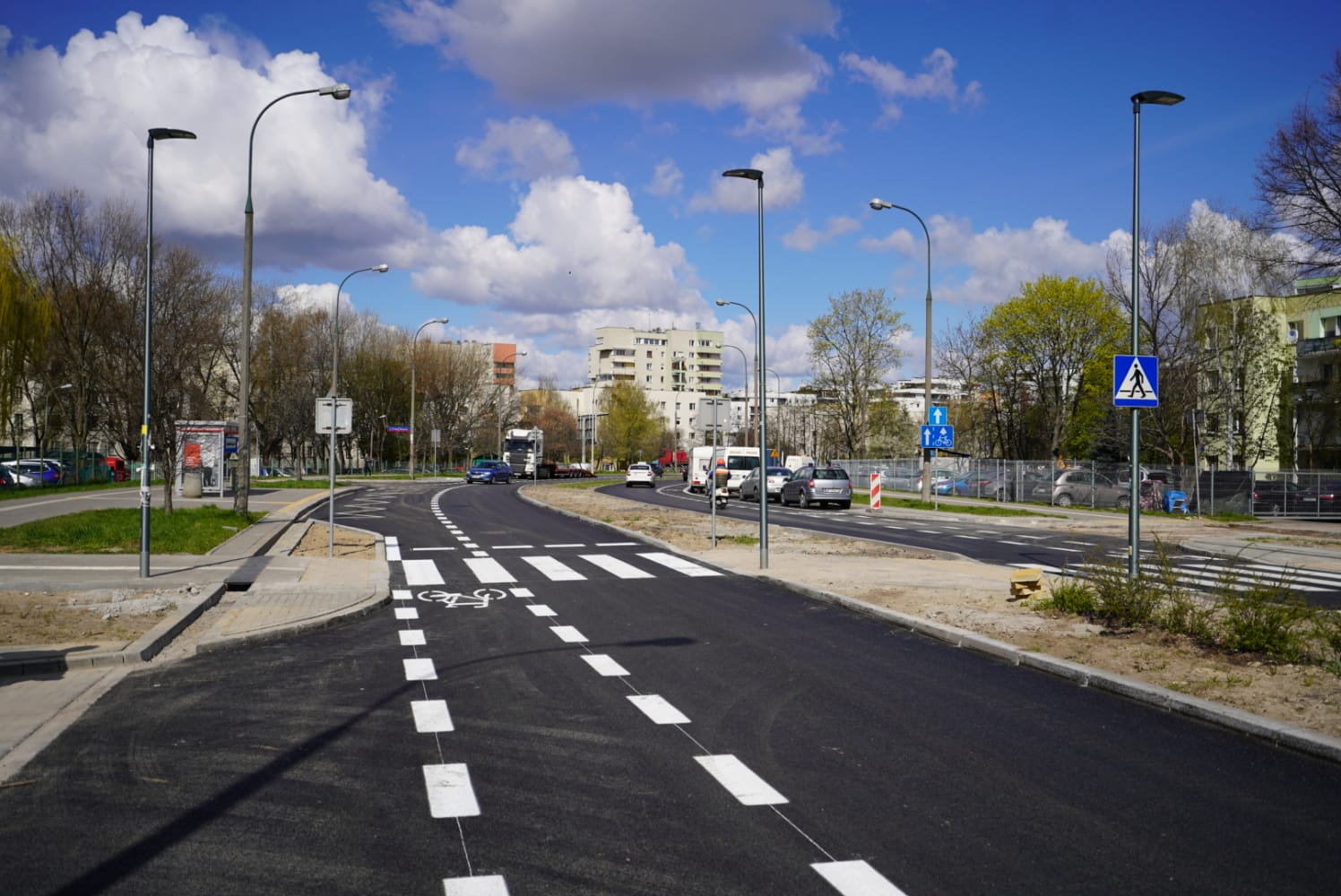 Nowo wyasfaltowana ulica miejska ze ścieżką rowerową i przejściem dla pieszych, w tle nowoczesne budynki.
