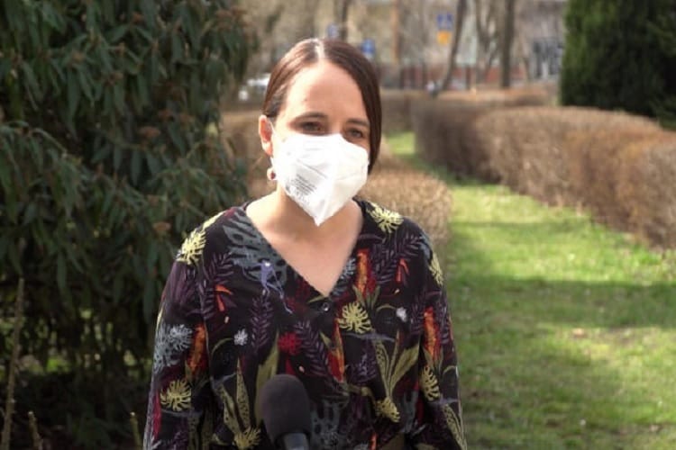 Kobieta ubrana w kwiecistą koszulę i maskę na twarzy przemawia do mikrofonu na zewnątrz.