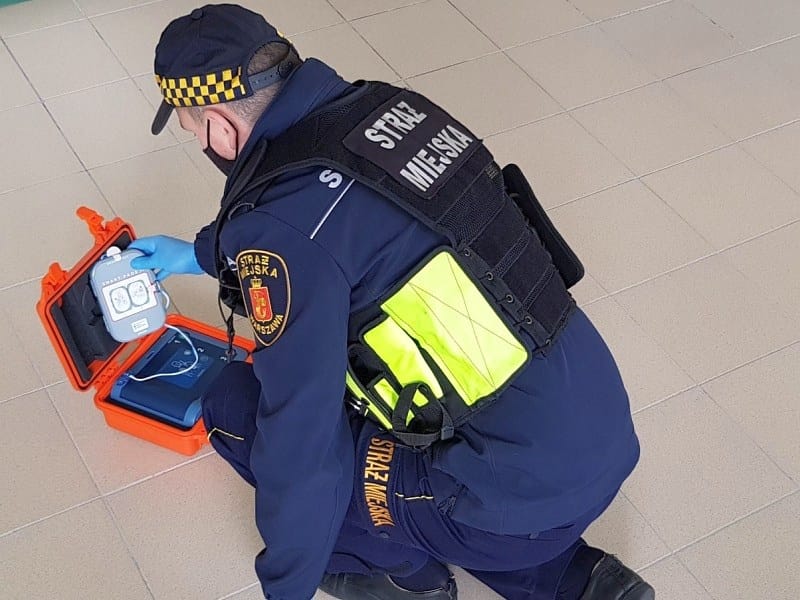 Strażnik miejski sprawdzający defibrylator AED. Na mundurze widoczny „Straż Miejski”.