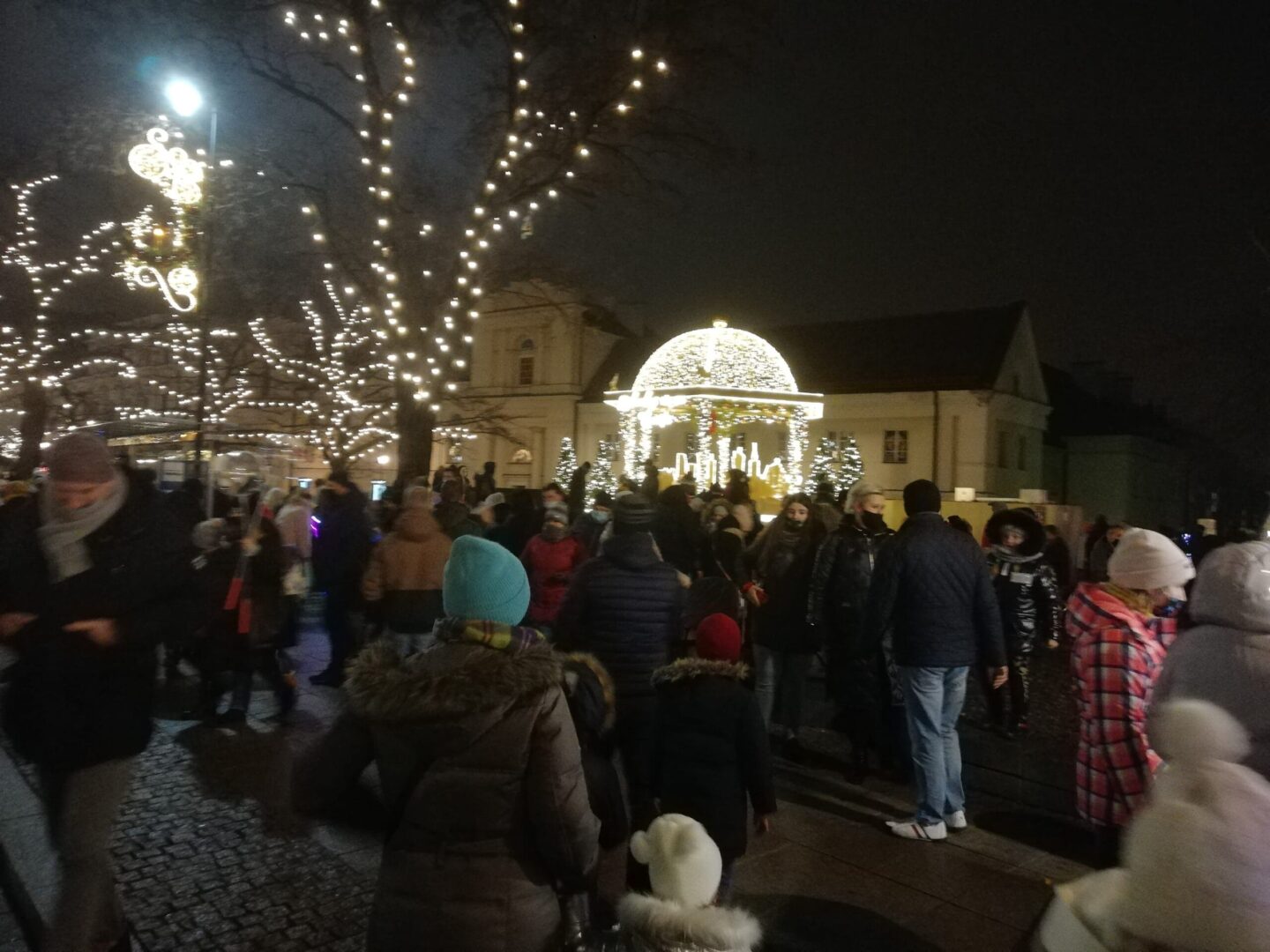 Nocny jarmark bożonarodzeniowy z altaną udekorowaną światłami i wieloma zgromadzonymi ludźmi.