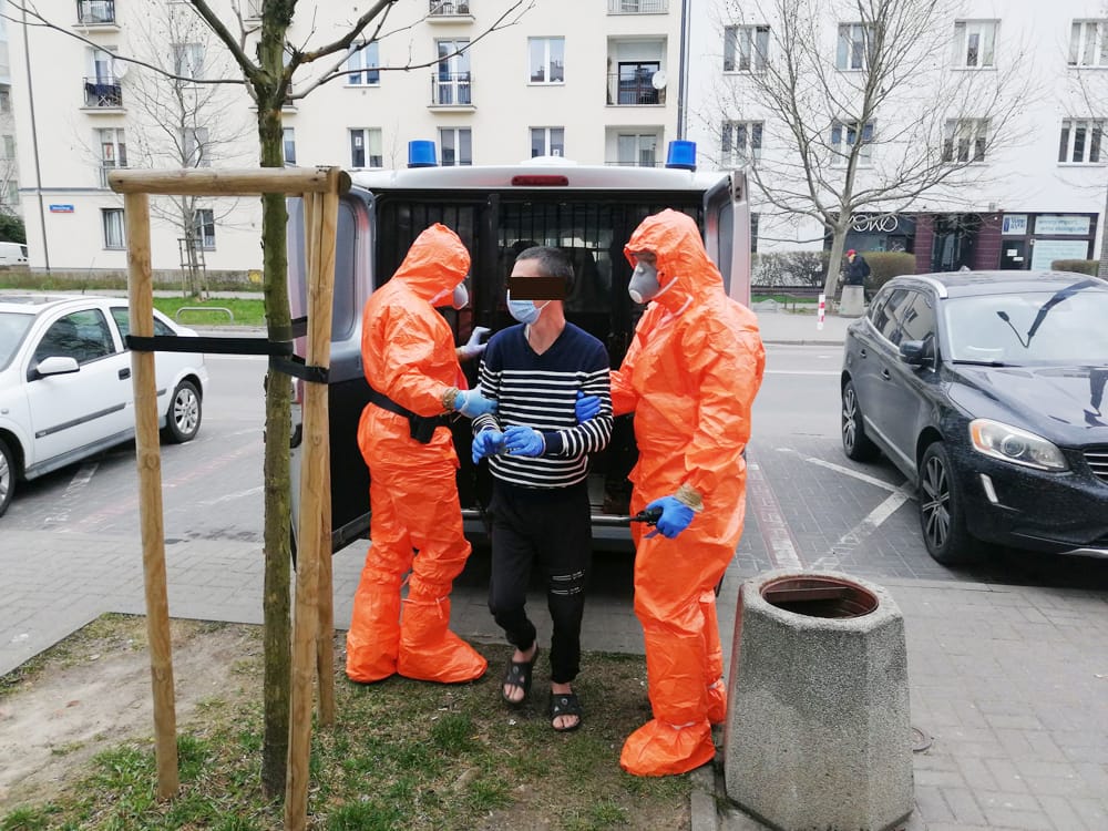 Dwóch pracowników służby zdrowia ubranych w kombinezony ochronne eskortuje osobę w kajdankach w pobliżu furgonetki.