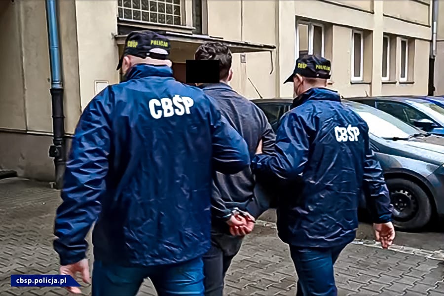 Policjanci eskortujący podejrzanego w kajdankach. Na kurtkach funkcjonariuszy widoczne są insygnia CBSP.