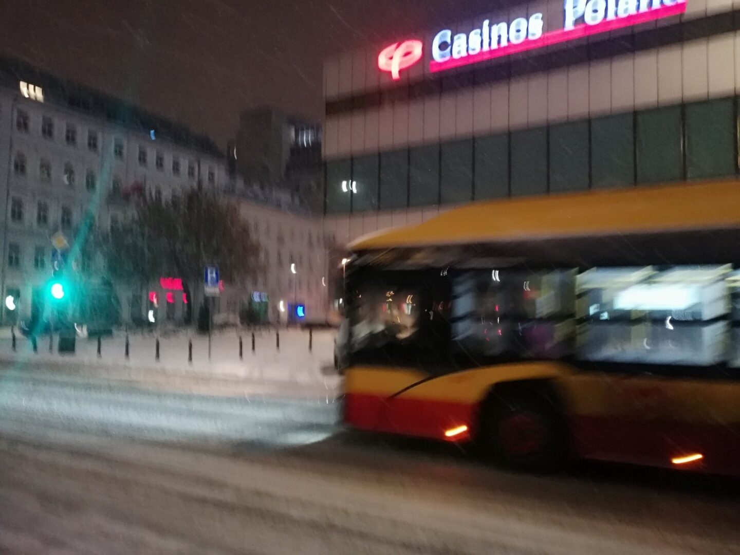 Rozmazany obraz autobusu jadącego nocą przez zaśnieżoną ulicę miasta, mijając budynek Casinos Poland.
