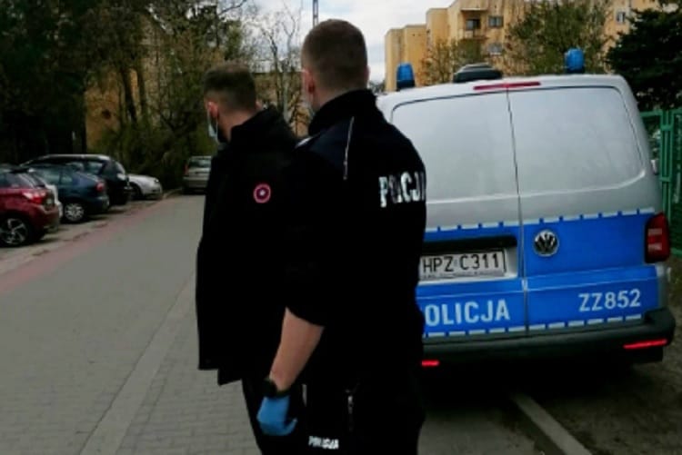 Dwóch polskich policjantów stoi przy radiowozie, jeden z nich ma na twarzy maskę.
