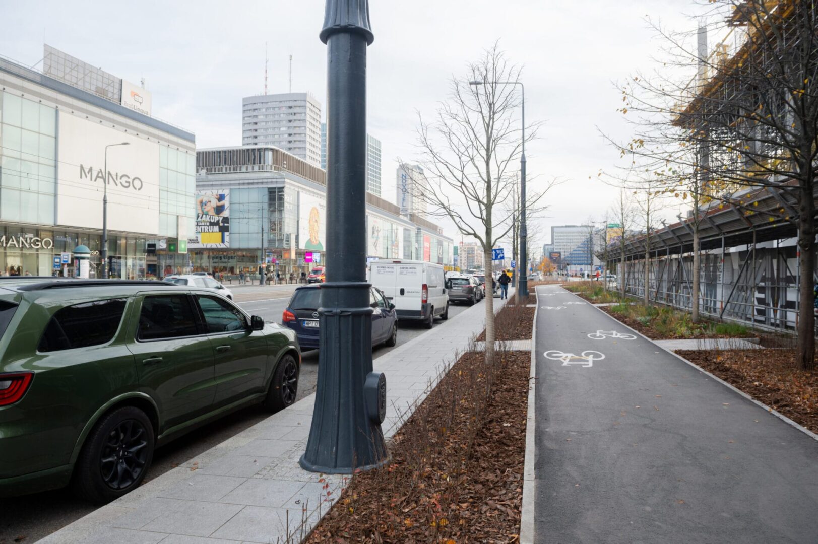 Nowoczesna ulica miejska z pasem rowerowym, sklepami i zaparkowanymi samochodami. Widoczny sklep „MANGO”.