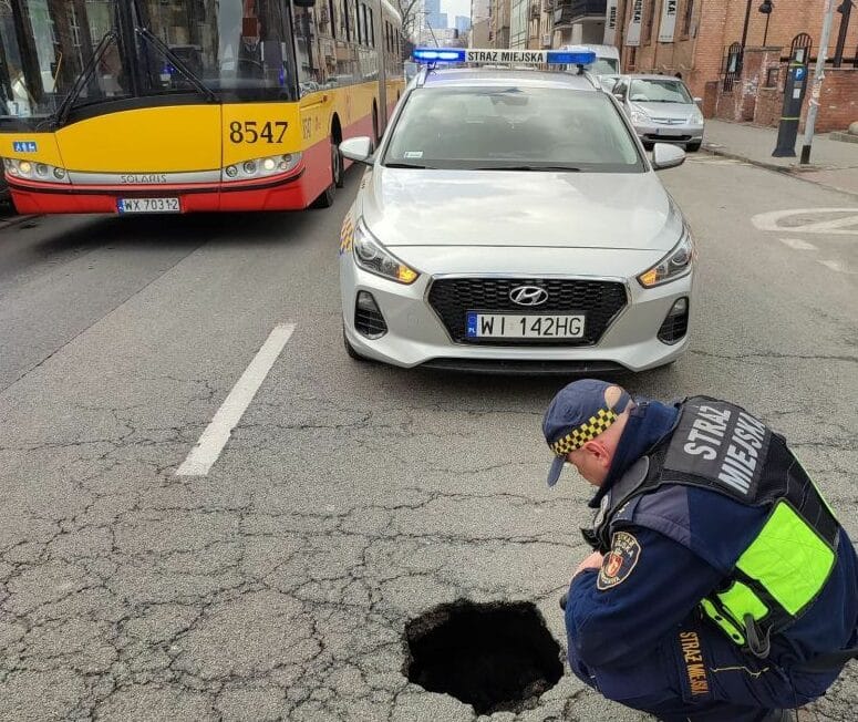 Inspektor miejski bada dziurę w drodze w pobliżu autobusu i radiowozu. „WI 142HG