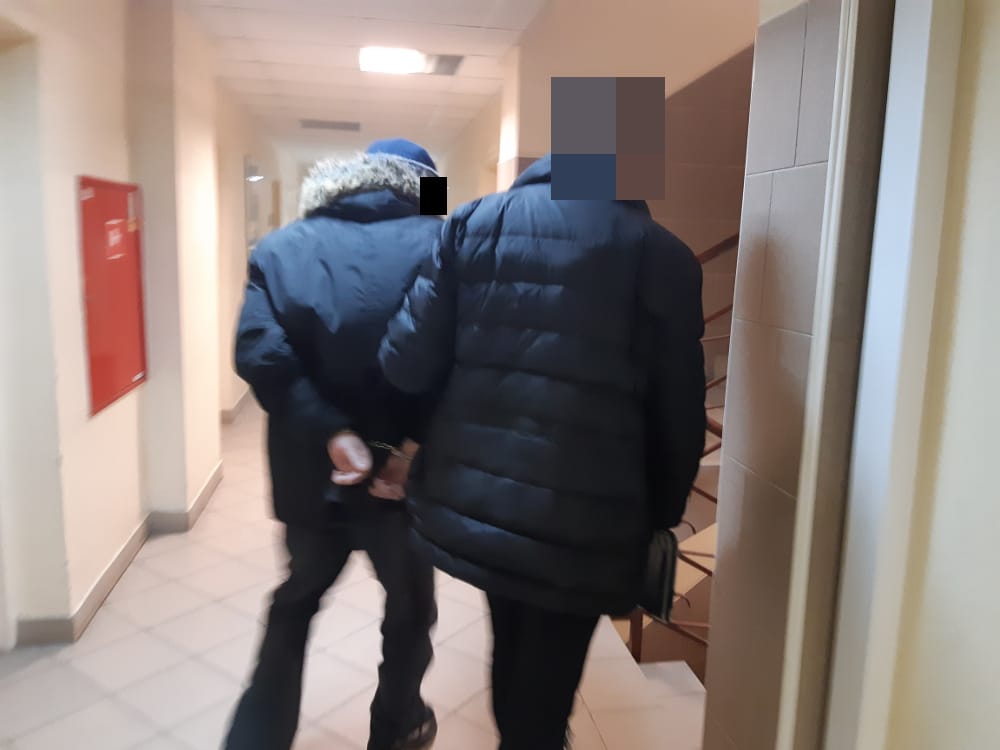 Dwie osoby w kajdankach idą korytarzem, eskortowane przez funkcjonariuszy organów ścigania.