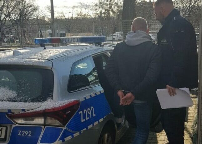 Dwóch mężczyzn w kajdankach stoi obok radiowozu. Z292