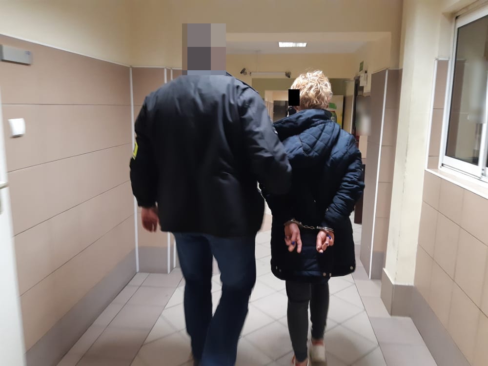 Policjant eskortujący skutą kajdankami kobietę korytarzem.