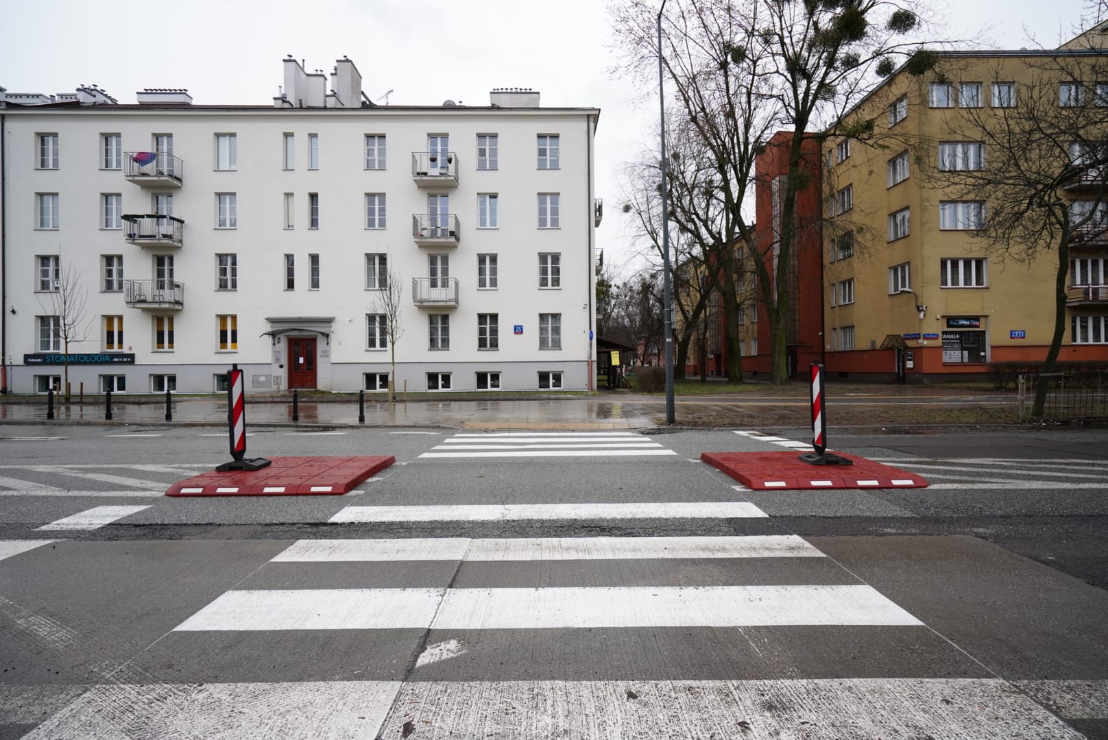 Przejście dla pieszych oznaczone czerwonymi i białymi słupkami przed budynkami mieszkalnymi.