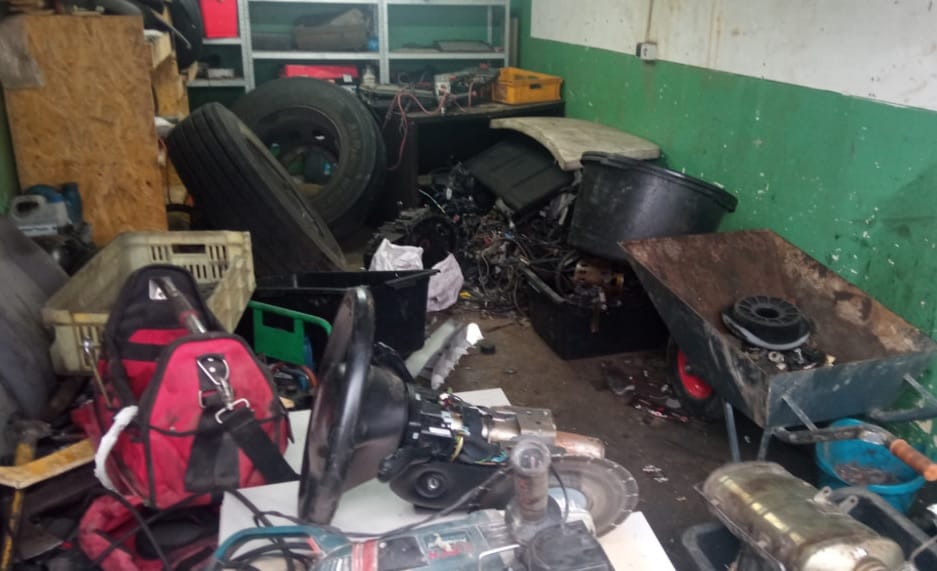 Brudny warsztat samochodowy z narzędziami, oponami i częściami samochodowymi porozrzucanymi na podłodze.