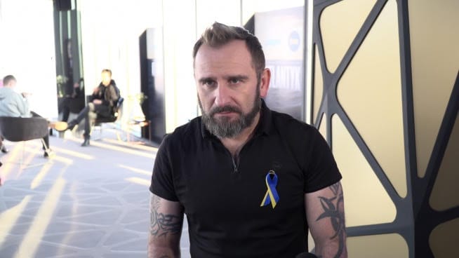 Mężczyzna z brodą i tatuażami, ubrany w czarną koszulę z ukraińską wstążką.