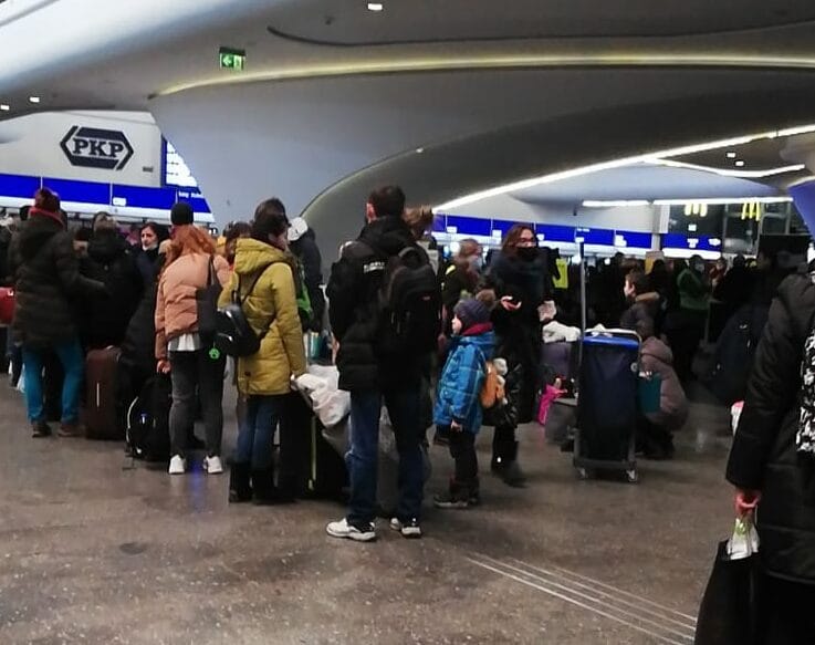 Zatłoczony dworzec kolejowy, pasażerowie czekają przy kasach biletowych; widoczne logo PKP.