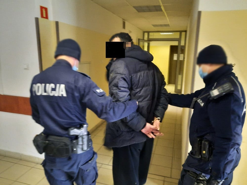 Policjanci eskortują skutego kajdankami podejrzanego korytarzem.