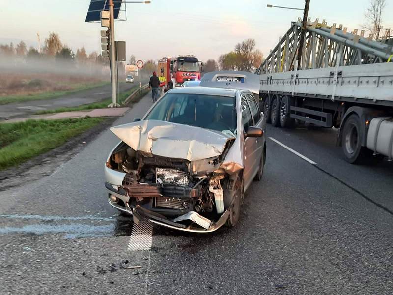 Uszkodzony srebrny samochód po zderzeniu z ciężarówką przewożącą metalową konstrukcję.